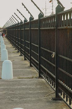 A perspective view of an iron fence with a clear sky, featuring concrete bollards along the path.